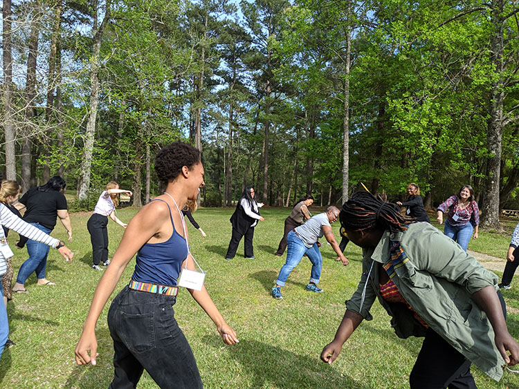 A multiracial group of leaders smiling and laughing as they stand in a circle outside on a lush green lawn surrounded by trees. They're swinging their arms and enjoying moving. 