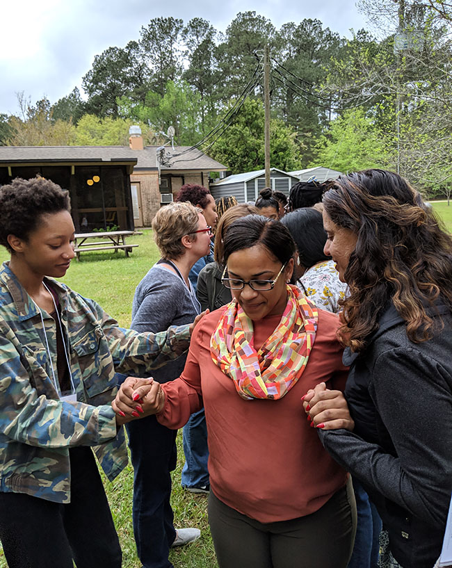 A woman with brown skin makes her way through way down the center of a group of people. Two people on either side of her are gently supporting her and offering affirming words. 