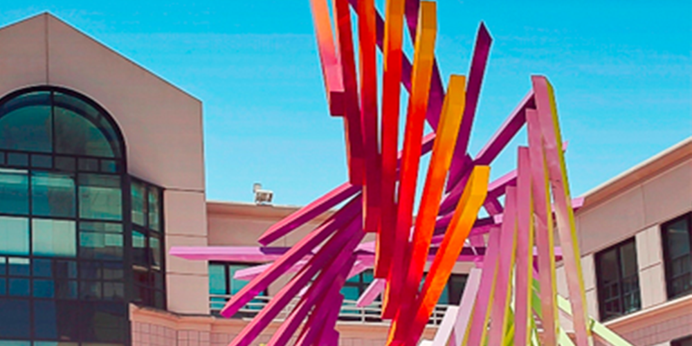 An exterior shot of 500 12th Street scene from the middle of 12 Street City Center. It is a beige building, with arch-shaped windows that reflect the sun. On the right, a yellow, orange and, purple abstract sculpture reaches up to the blue sky. 