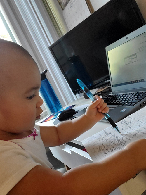 An adorable 15 month old "works" from home sitting at a desk with a pen in her hand.